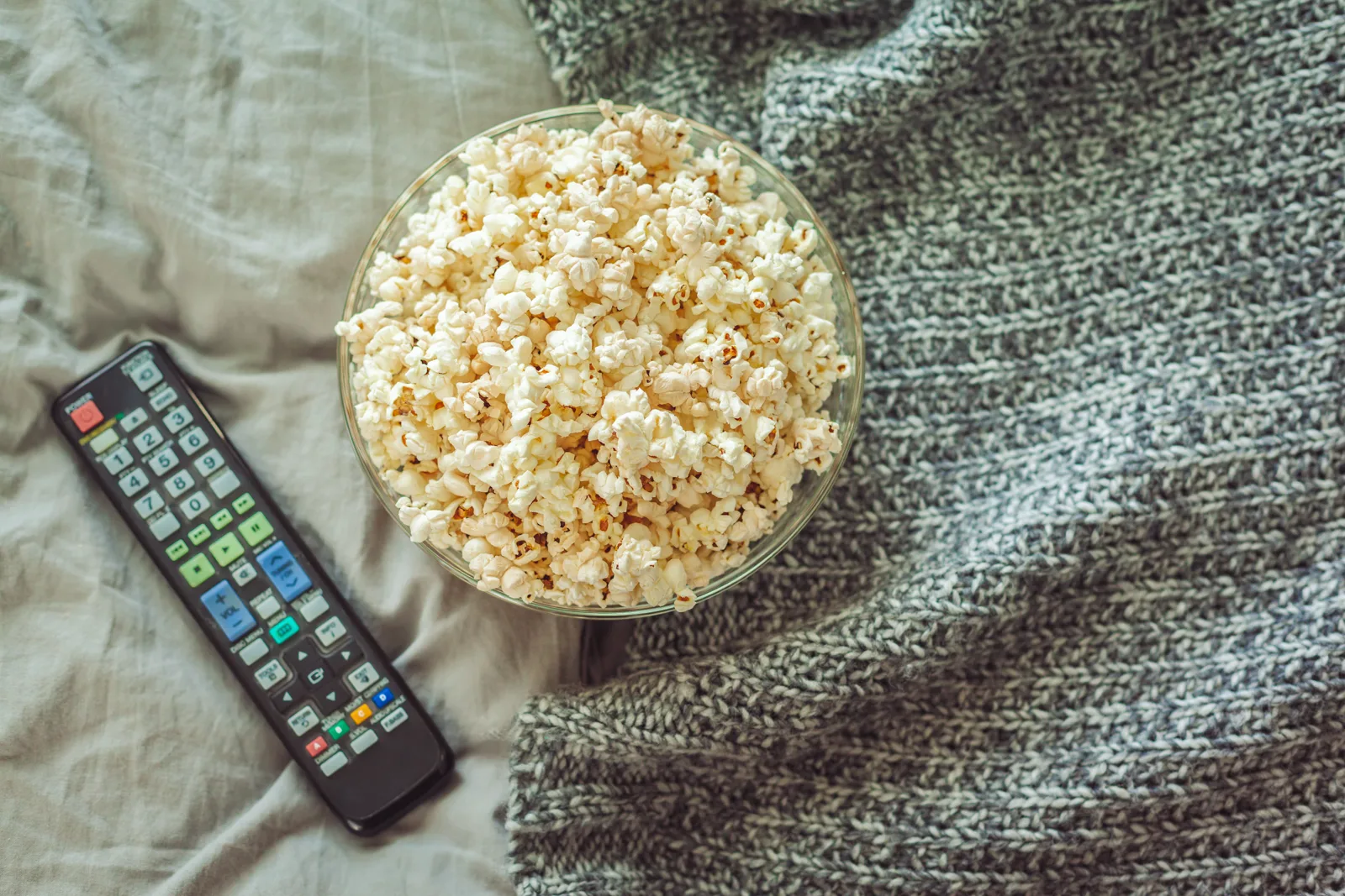 Bowl of popcorn and a cozy blanket on a couch in front of a glowing screen